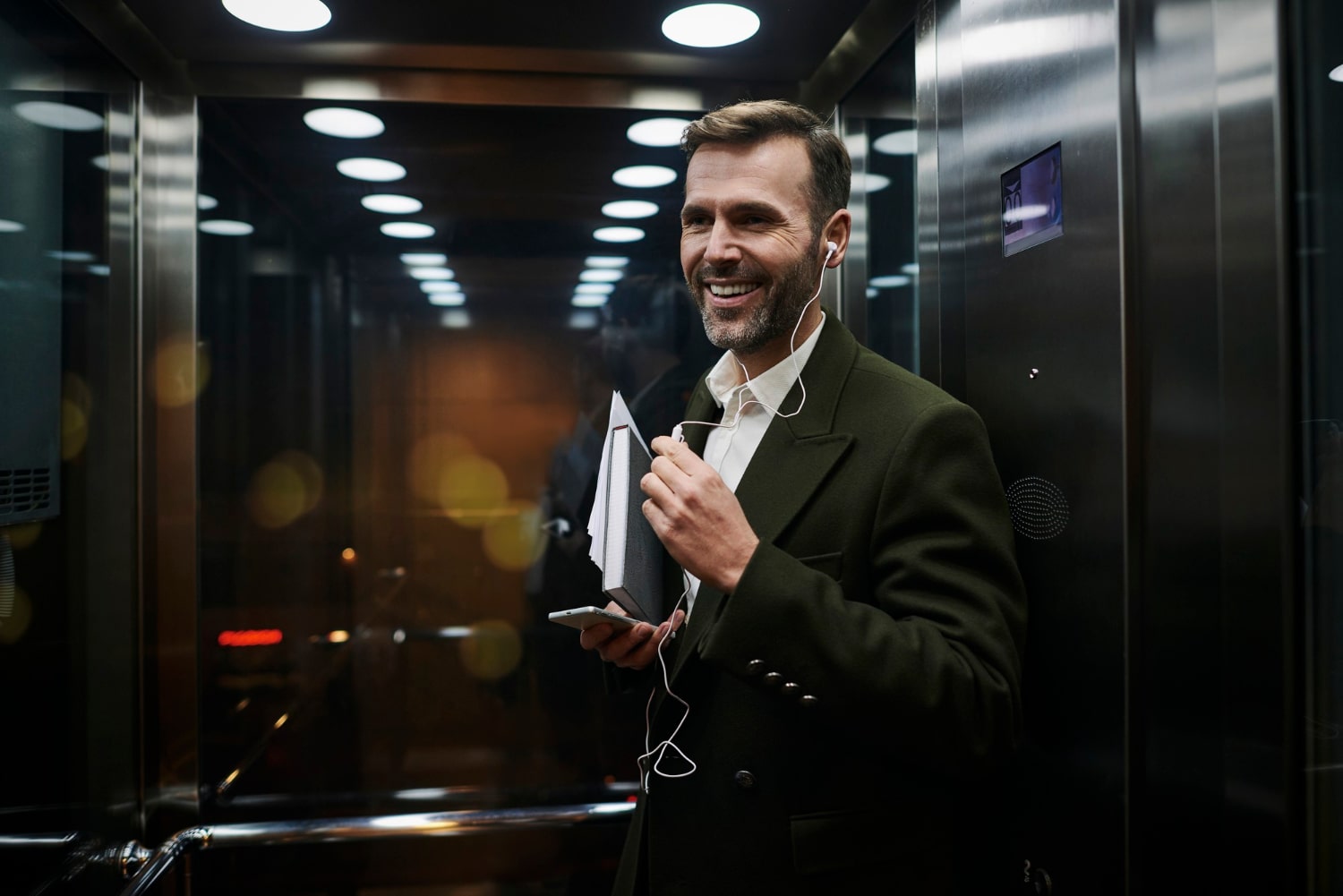 Man in suit smiling with headphones and holding a book in an elevator.