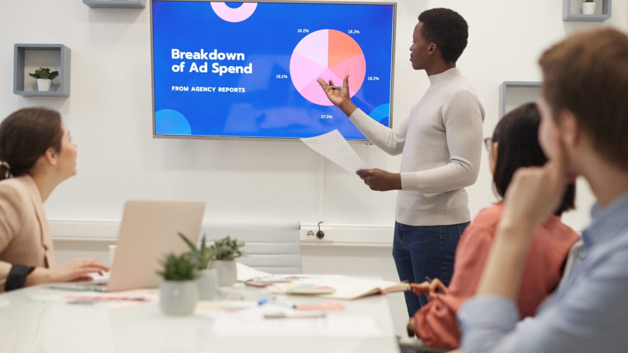 Person presenting ad spend breakdown on screen to group of colleagues in a meeting room.