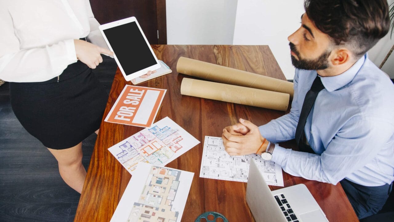 Real estate agents discussing floor plans and property details at a desk with a laptop and "For Sale" sign.