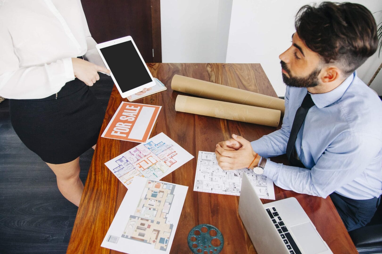 Real estate agents discussing floor plans and property details at a desk with a laptop and "For Sale" sign.
