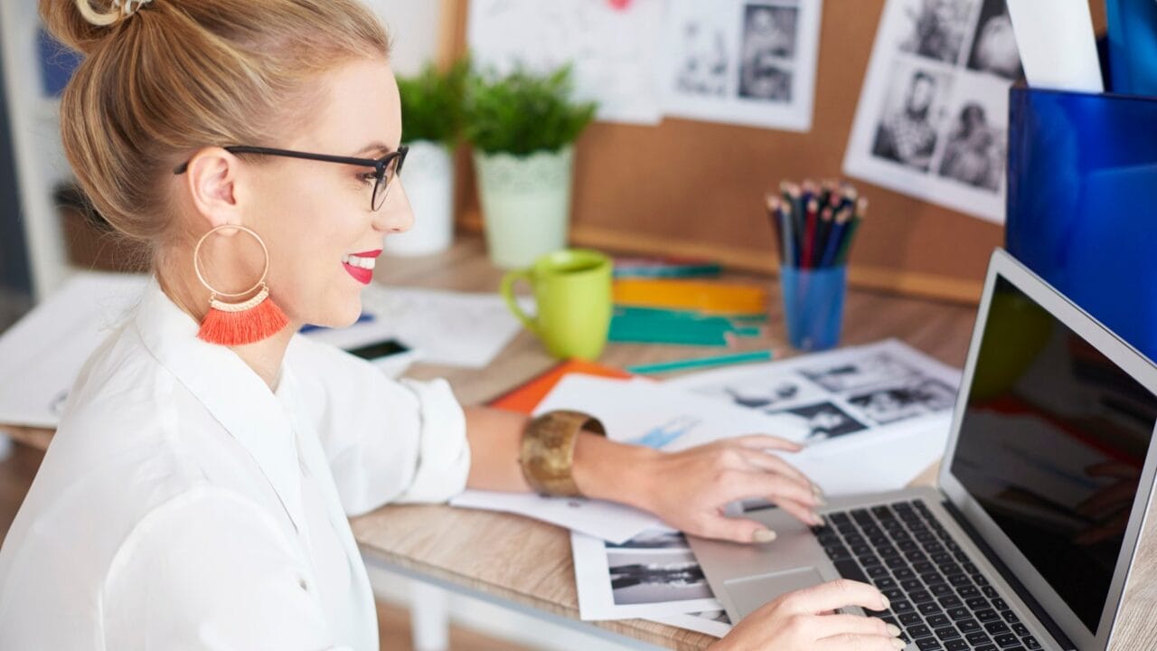 Woman working on a laptop at a desk, surrounded by papers and office supplies.