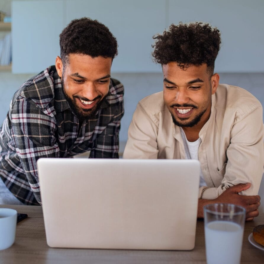 Two people smiling and looking at a laptop screen in a cozy room with glasses of water on the table.