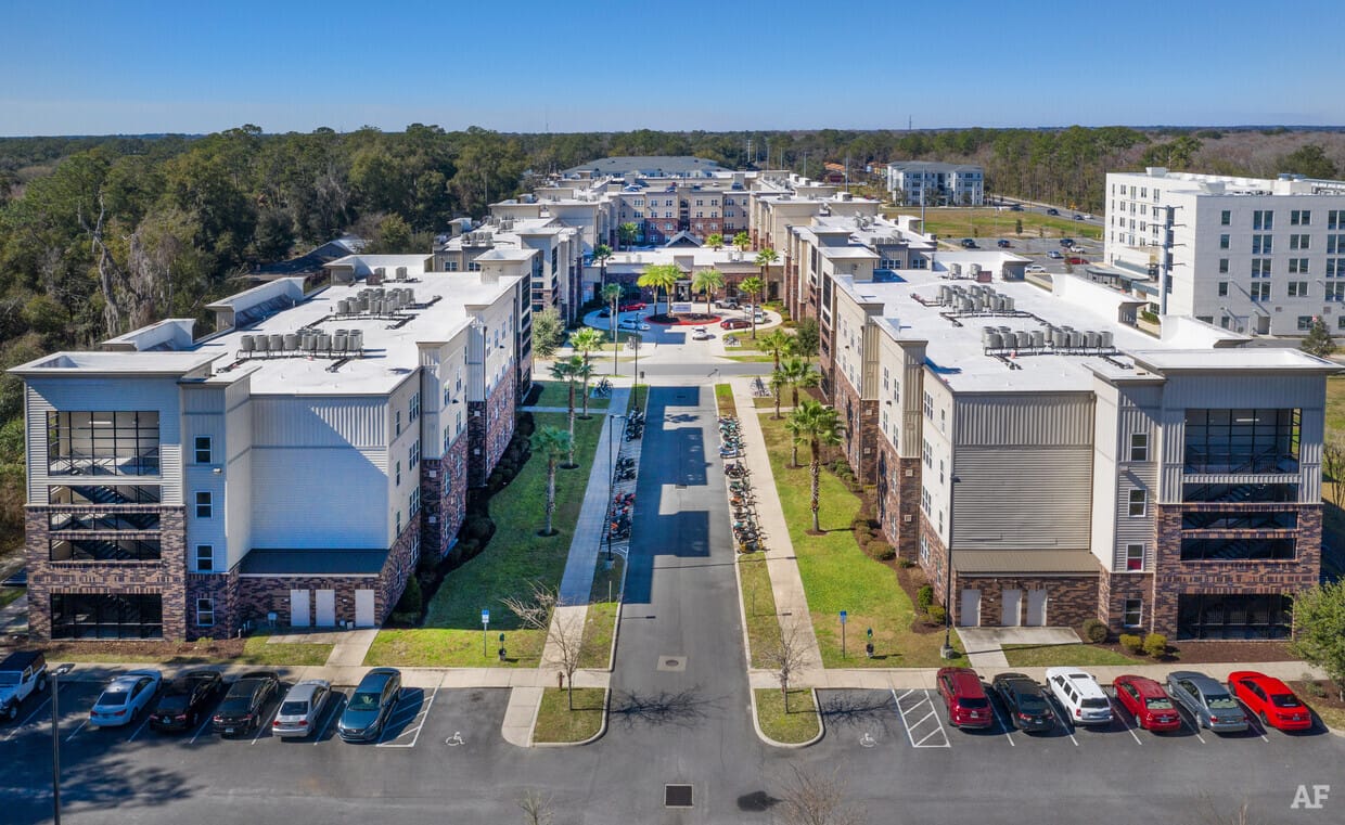 Aerial view of a modern apartment complex with parked cars, landscaped entrance, and surrounding greenery.