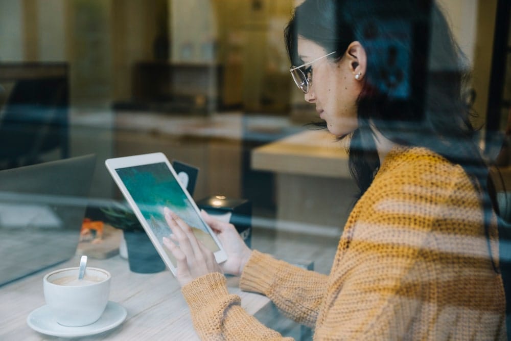 Person in a cafe using a tablet, wearing a yellow sweater, with a cup of coffee beside them.