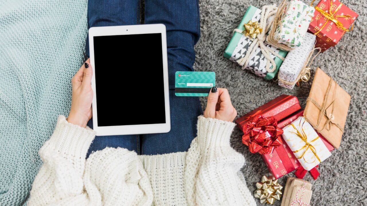 Person holding tablet and credit card, sitting on carpet with wrapped gifts nearby, suggesting online holiday shopping.