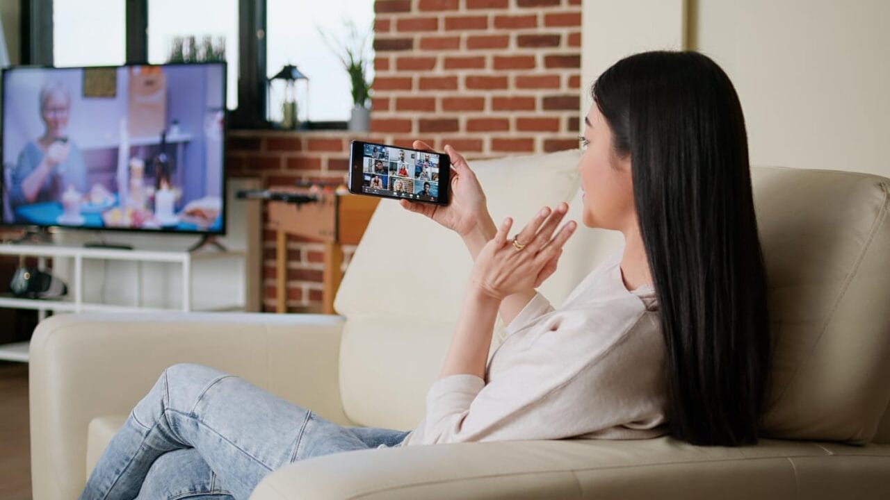 Woman on a couch video calling on smartphone with TV in the background.