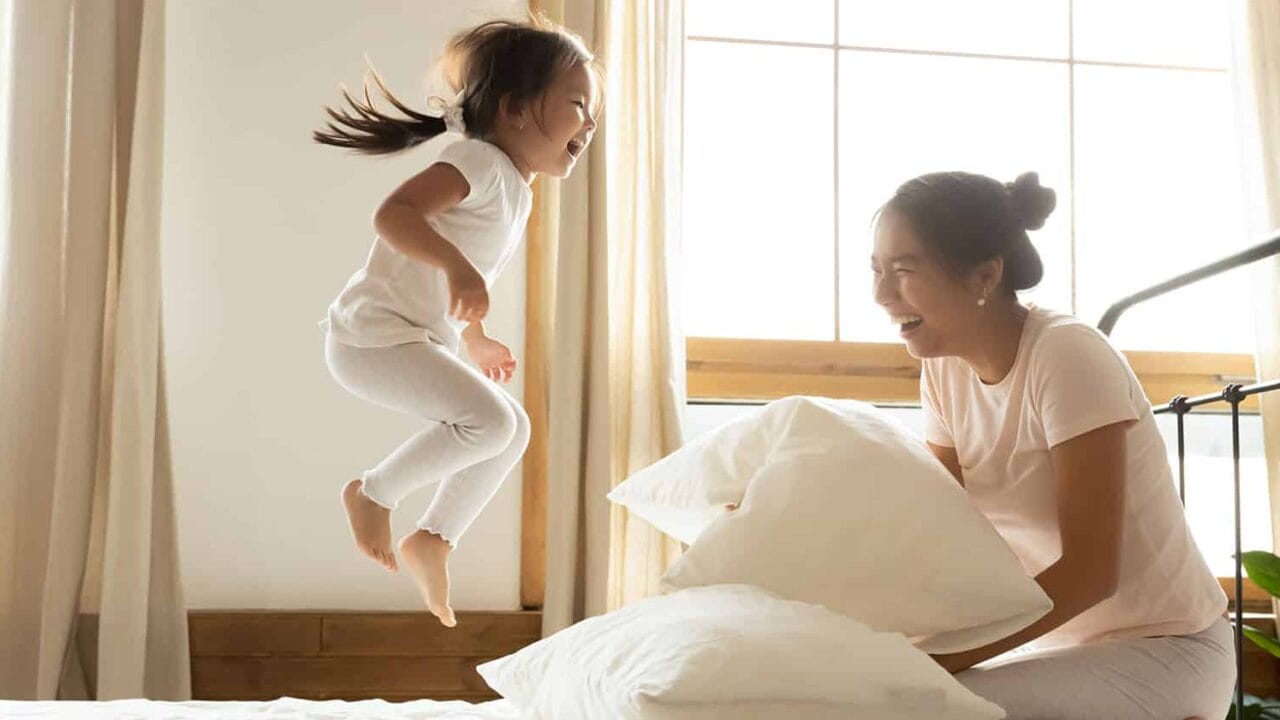 Child joyfully jumping on bed while mother holds pillow, both laughing in a sunlit bedroom with large windows and curtains.