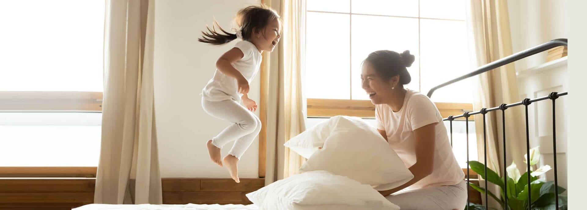 Child joyfully jumping on bed while mother holds pillow, both laughing in a sunlit bedroom with large windows and curtains.