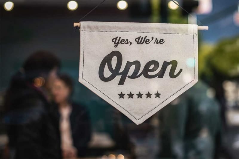 Sign in shop window reads "Yes, We're Open" with five stars underneath, blurred people in the background.