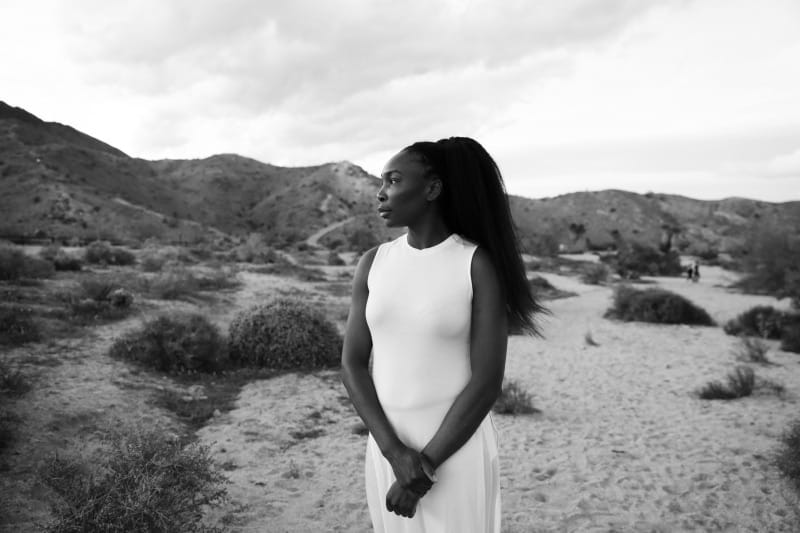 Person in a white dress standing in a desert landscape with mountains in the background, looking into the distance.