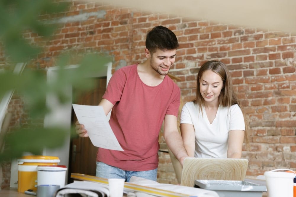 Two people collaborating on a project with a laptop and documents, in a workspace with a brick wall background.