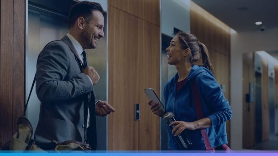 Man in suit and woman in athletic wear chatting near an office elevator, both smiling and holding bags.