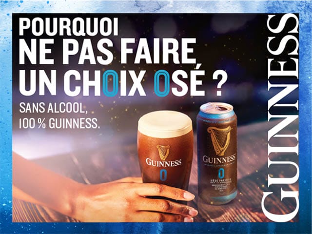 Hand holding a pint of non-alcoholic Guinness beer beside a can on a wooden table with promotional text in French.