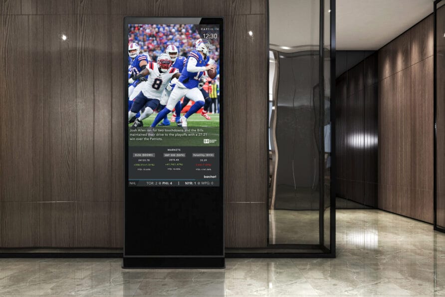 Digital kiosk displaying a football game broadcast with players in action, located in a modern building lobby.