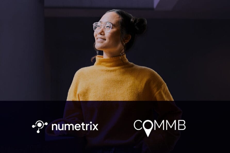 Person in a yellow sweater with glasses stands smiling in a dimly lit room, logos for numetrix and COMM below.