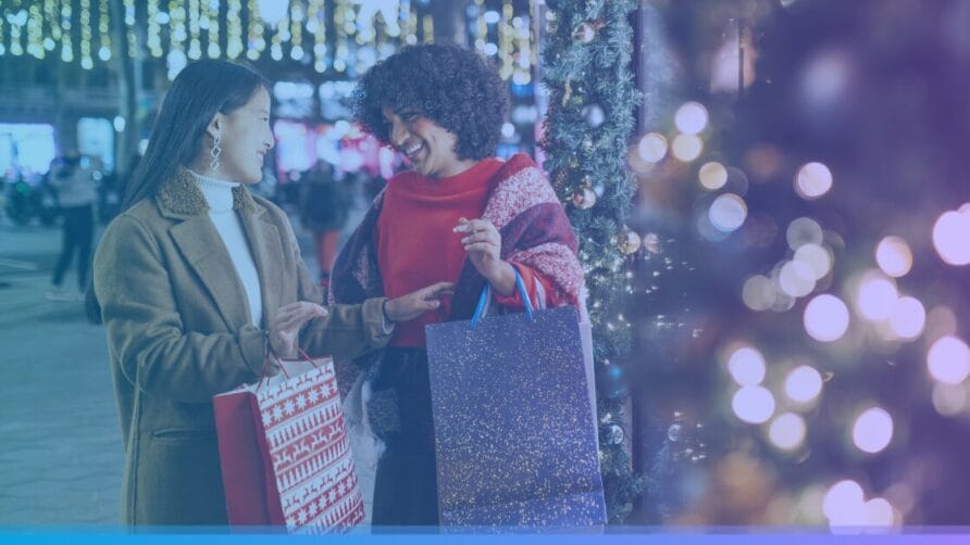 Two women smiling with shopping bags, standing near a decorated Christmas tree with festive lights in the background.